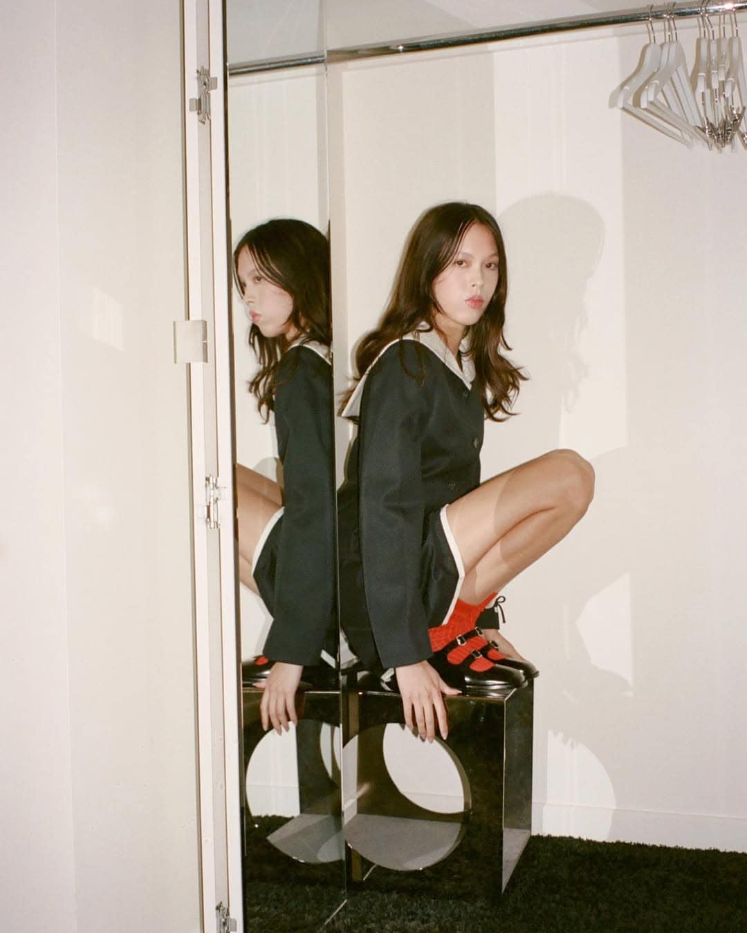 Model squatting on a reflective stool in a dark sailor-style dress with white trim and red socks against a light wall
