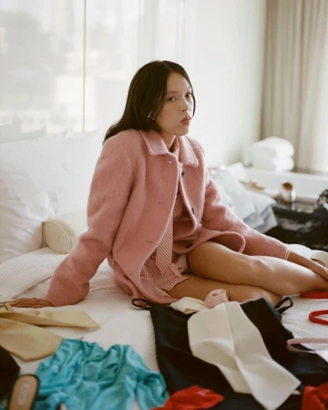 Model seated on a bed wearing a pink coat with various clothing items scattered around her in a hotel room setting