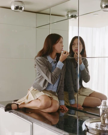 Model applying lipstick while sitting on a ledge in front of a mirrored wall, wearing a grey cardigan and pleated skirt.