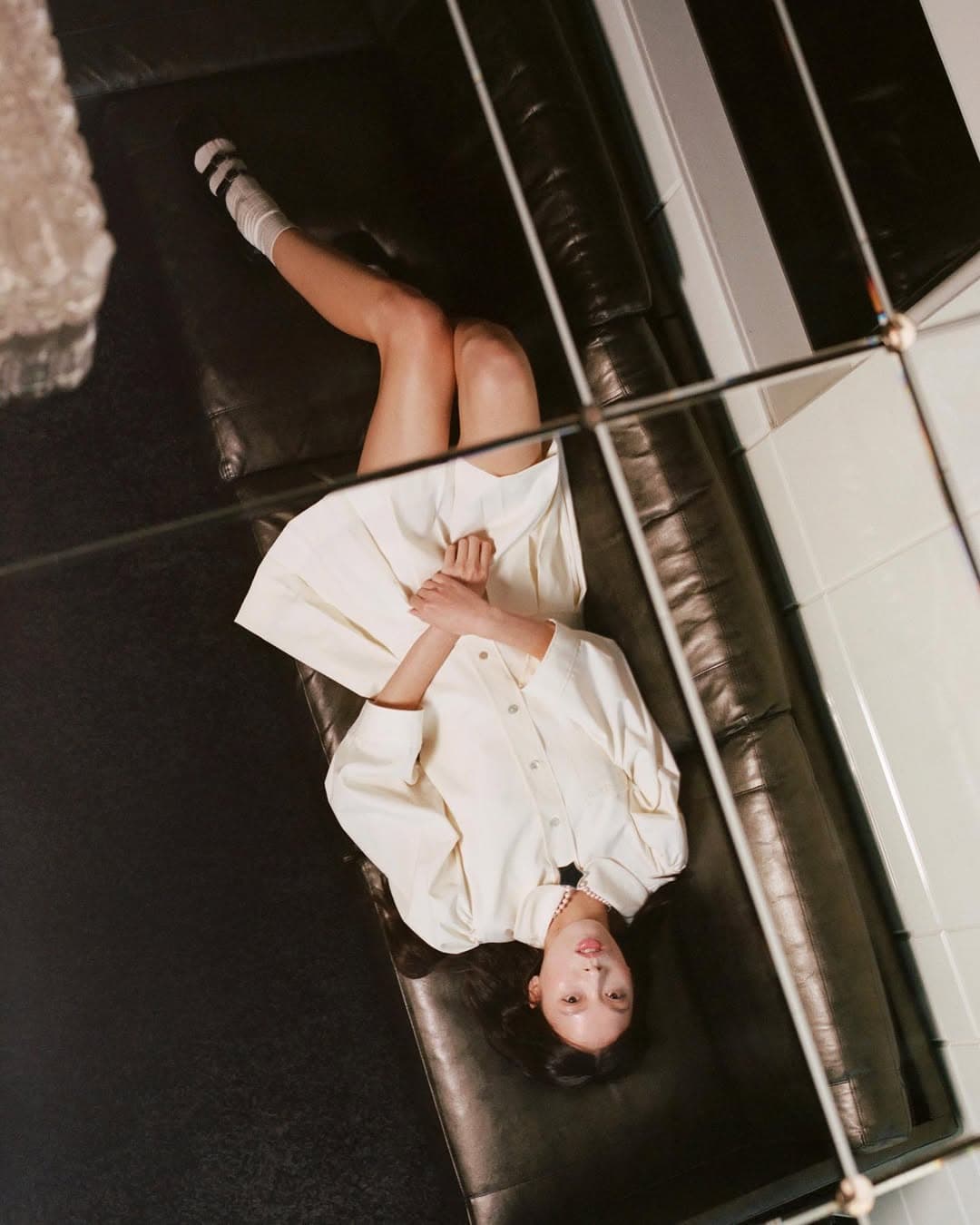 Model lying on a black leather sofa wearing a white dress and socks, viewed from an overhead angle behind a grid mirror.