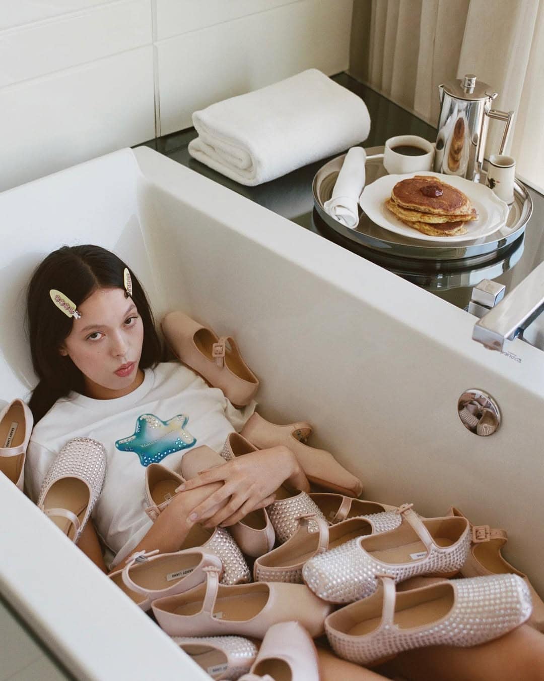 Model lying in a white bathtub surrounded by multiple pairs of pink shoes with a tray of pancakes nearby.