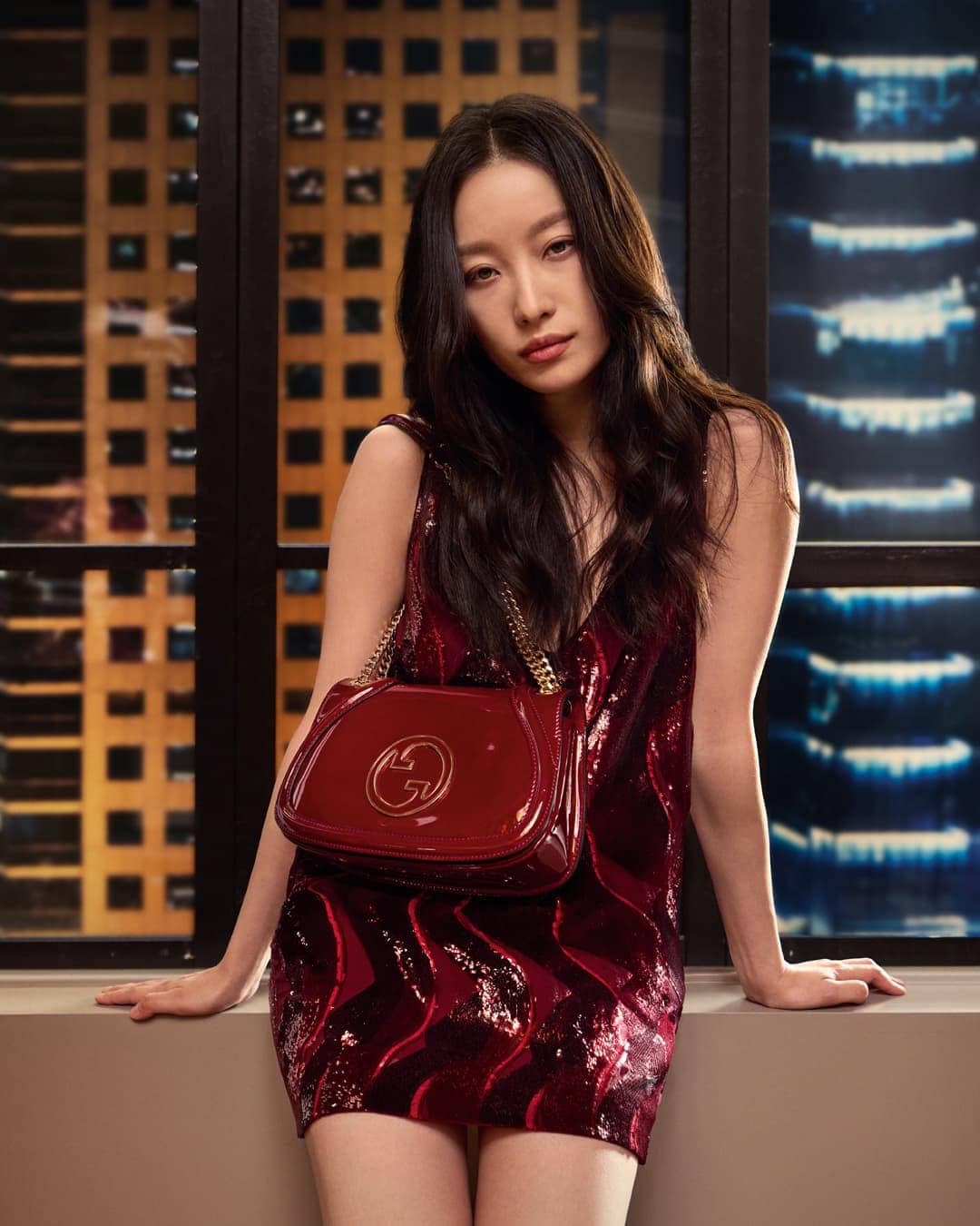 Ni Ni posing in a red sequin dress holding a matching leather handbag against a window with lit city buildings at night.