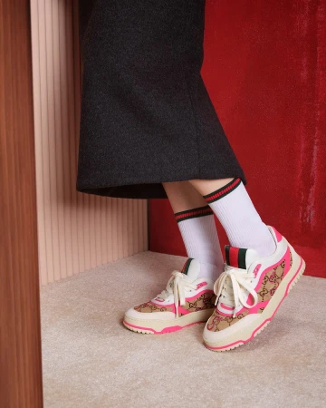 Close-up of a person wearing Gucci sneakers with white socks and a dark skirt against a red and ribbed textured backdrop