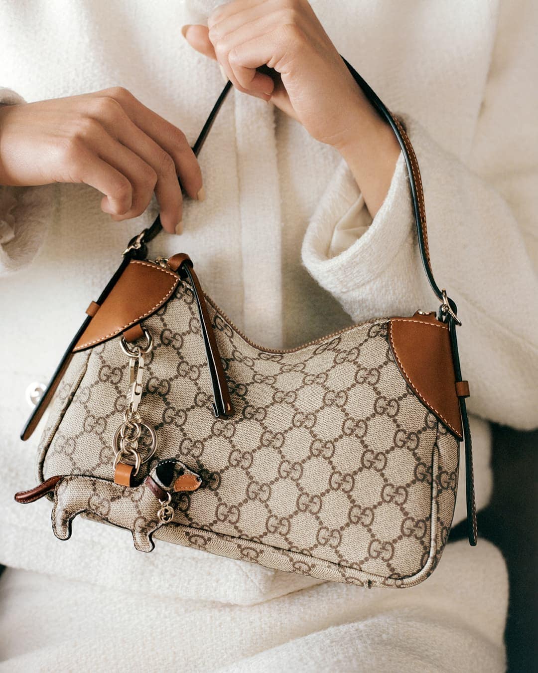 Person holding a monogram canvas handbag with brown leather trim and a dachshund bag charm against a white textured coat