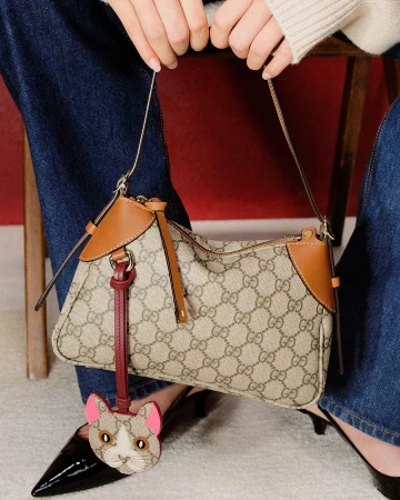Close-up of a person holding a monogram canvas shoulder bag with a cat charm, wearing blue jeans and black shoes against a red and beige backdrop