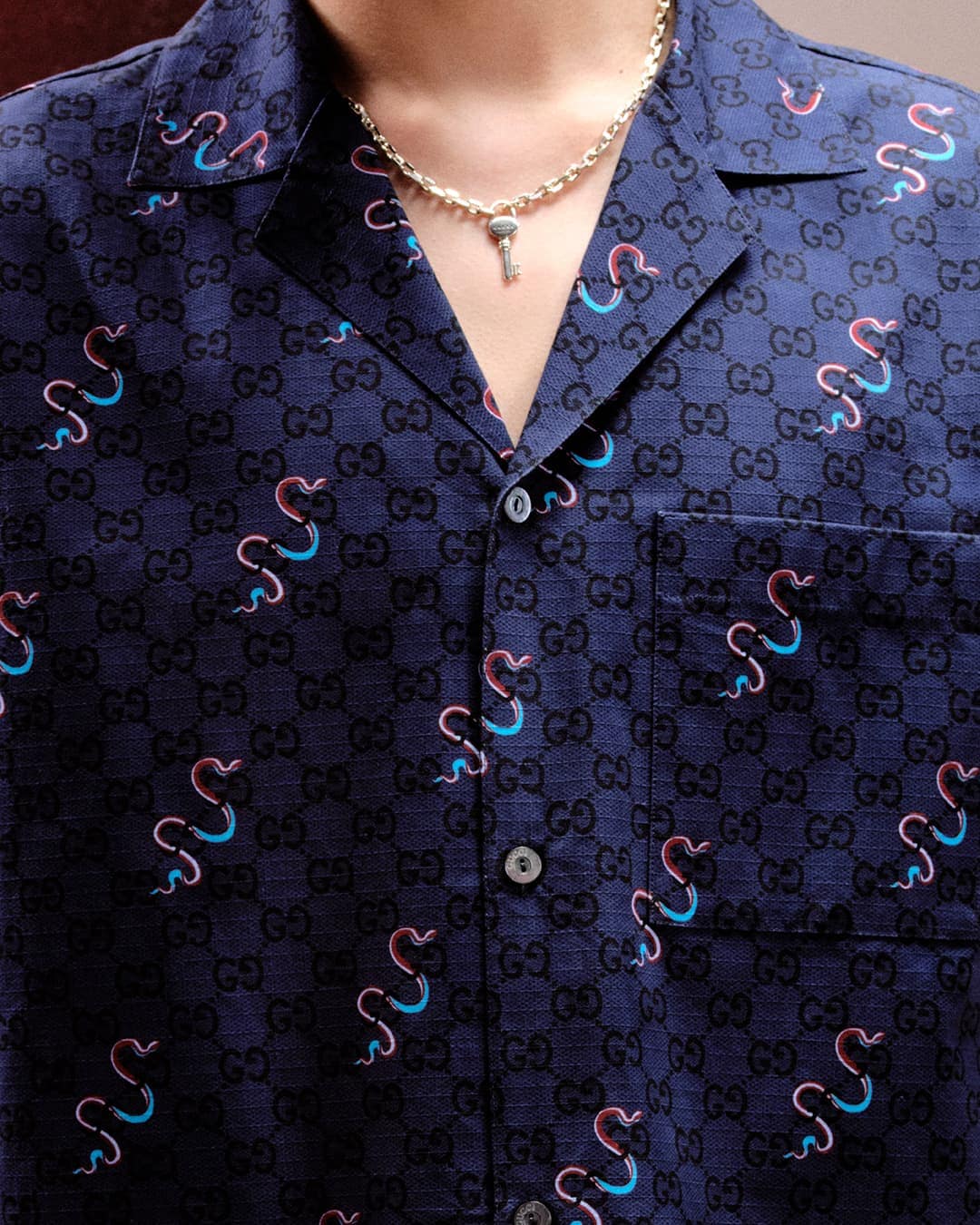 Close-up of a person wearing a navy blue Gucci patterned shirt with snake motifs and a gold key necklace.