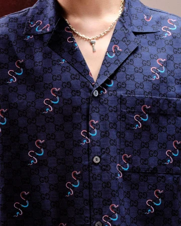 Close-up of a person wearing a navy blue Gucci patterned shirt with snake motifs and a gold key necklace.
