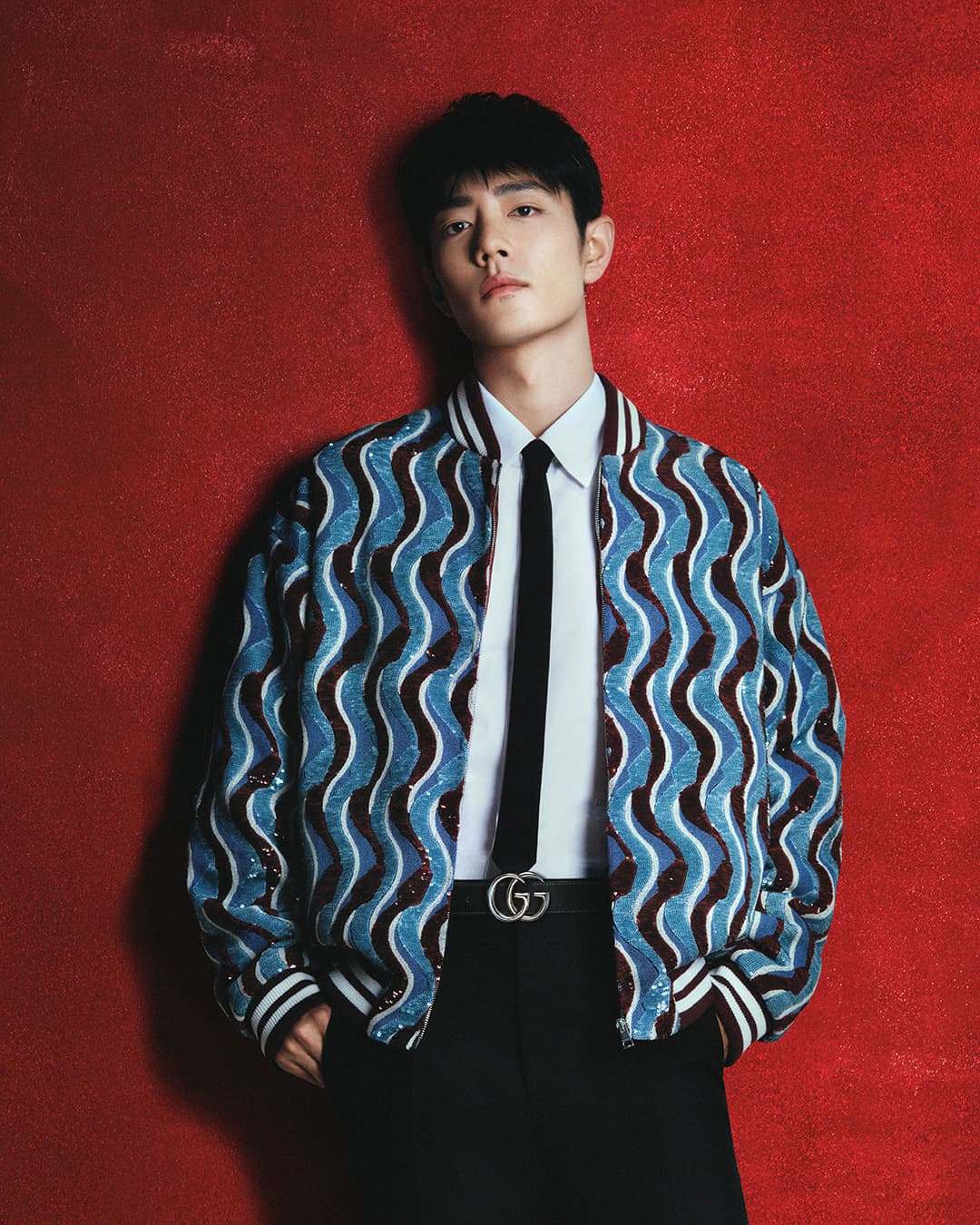 Portrait of Xiao Zhan wearing a patterned blue bomber jacket and black tie against a textured red background for Gucci.