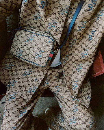 Close-up of a person wearing a monogram jacket and matching trousers with blue cross-body handbag by Gucci.