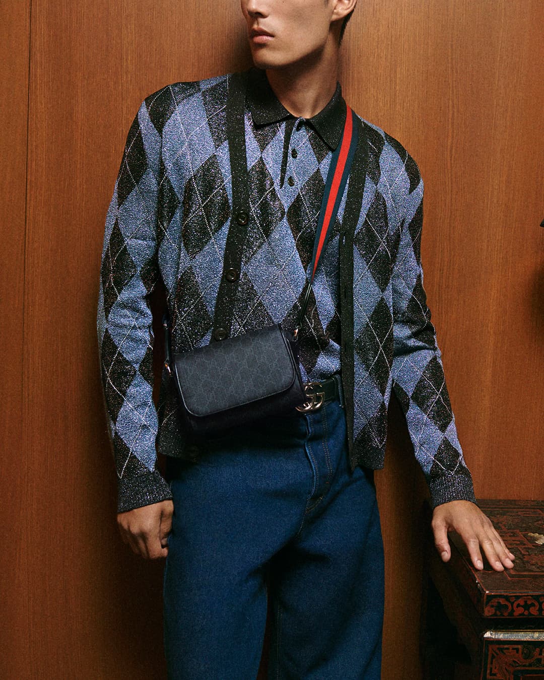Man wearing blue argyle polo cardigan and denim jeans standing against a wood wall for Gucci campaign