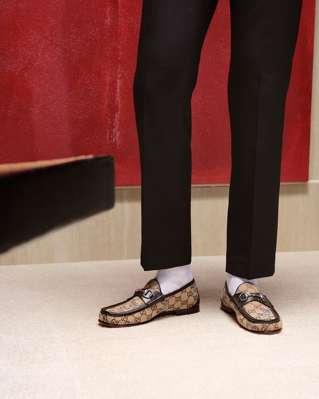 Close-up of a person wearing black trousers, white socks, and monogram horsebit loafers against a neutral and red background.
