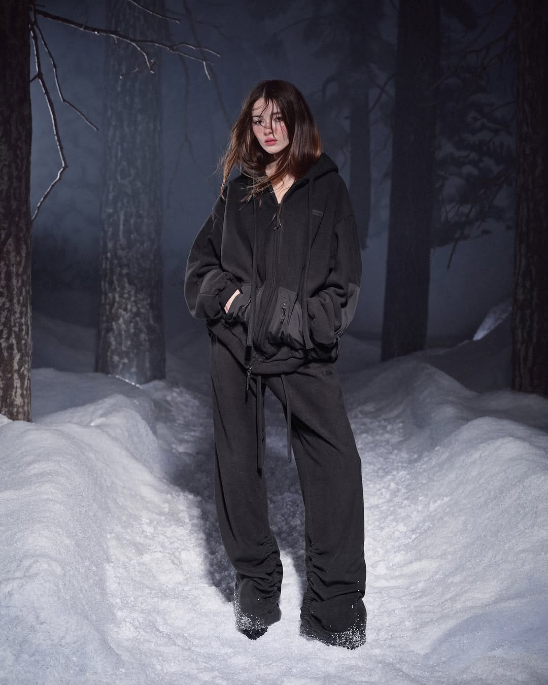 Model standing in a snowy forest wearing a black hoodie and matching trousers for the SKIMS Cold Weather campaign.