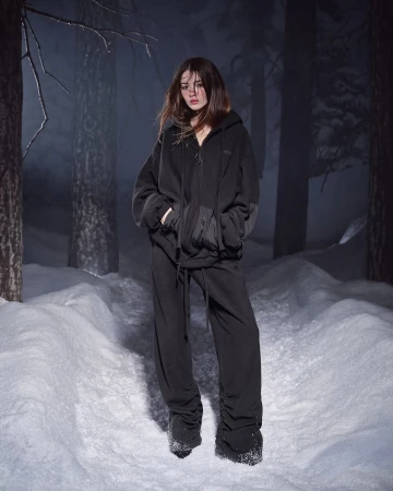 Model standing in a snowy forest wearing a black hoodie and matching trousers for the SKIMS Cold Weather campaign.