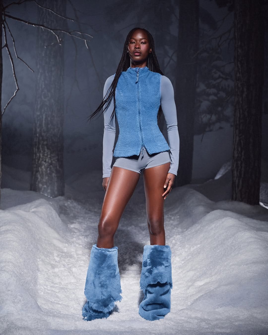 Model wearing blue fuzzy vest, matching furry boots, and grey shorts standing on a snowy forest path.