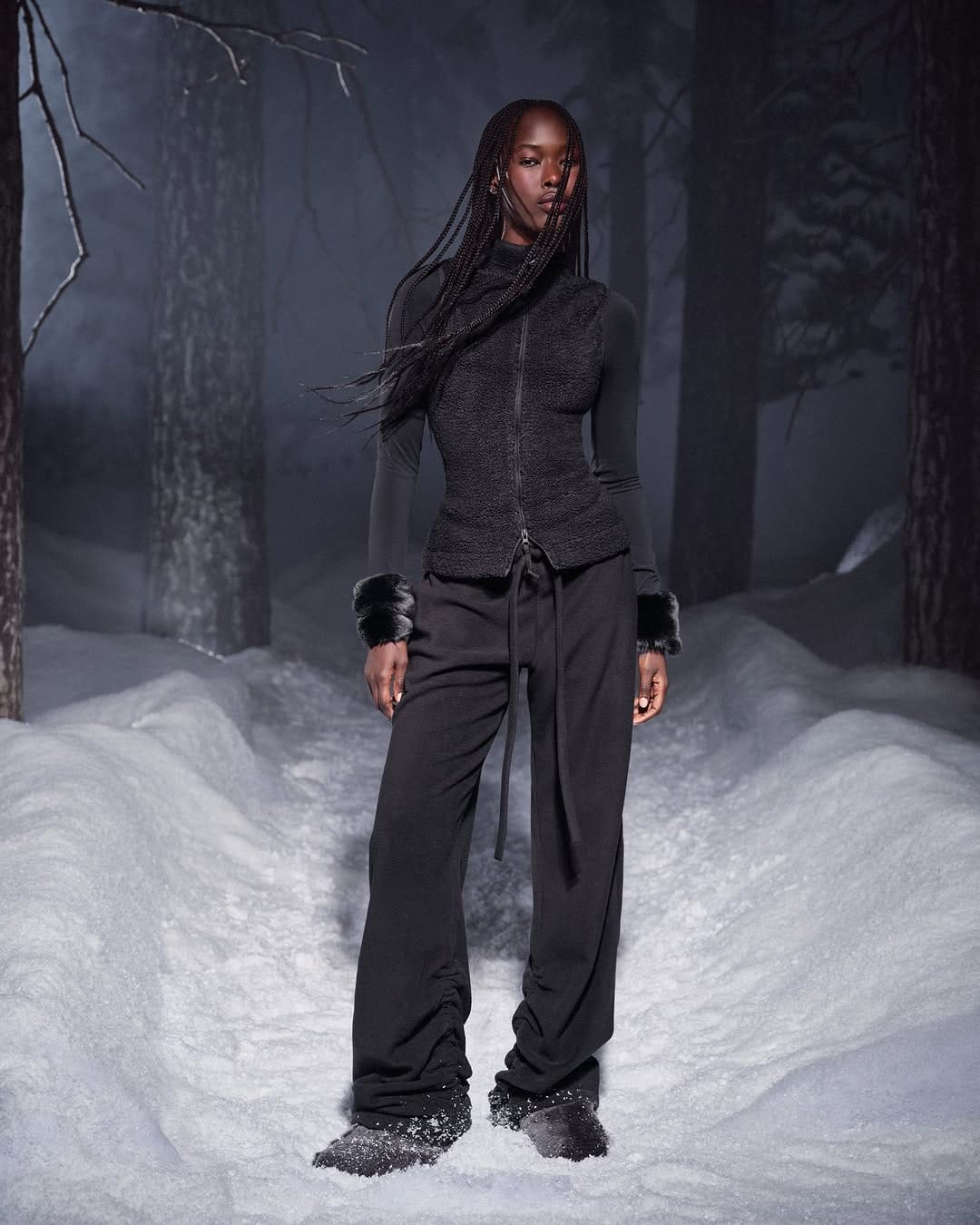 Model with long braids wearing a black zip vest and pants standing in a snowy forest at dusk