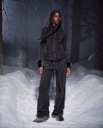 Model with long braids wearing a black zip vest and pants standing in a snowy forest at dusk