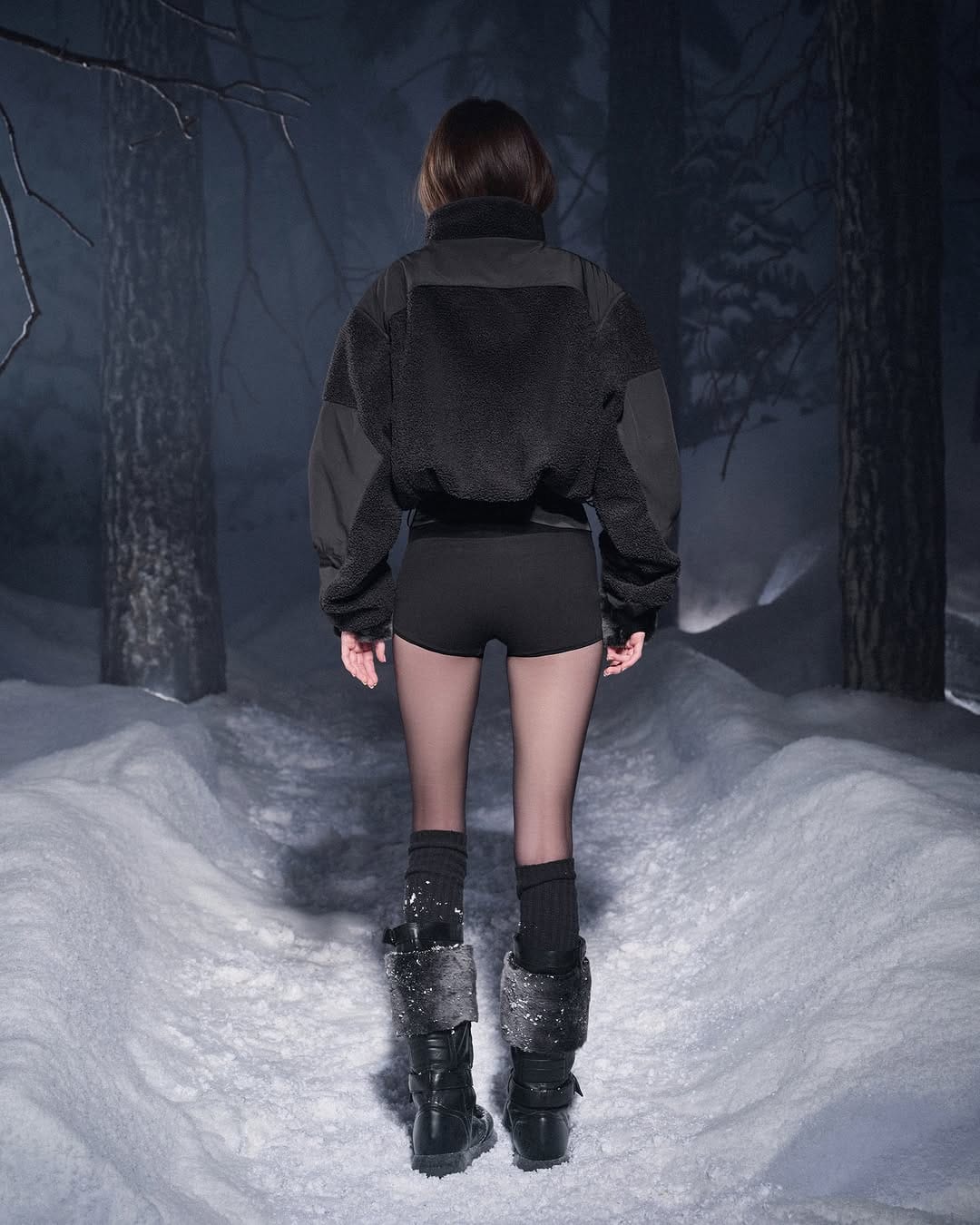 Model from behind wearing a black fleece jacket and winter boots on a snowy path in a forest at night