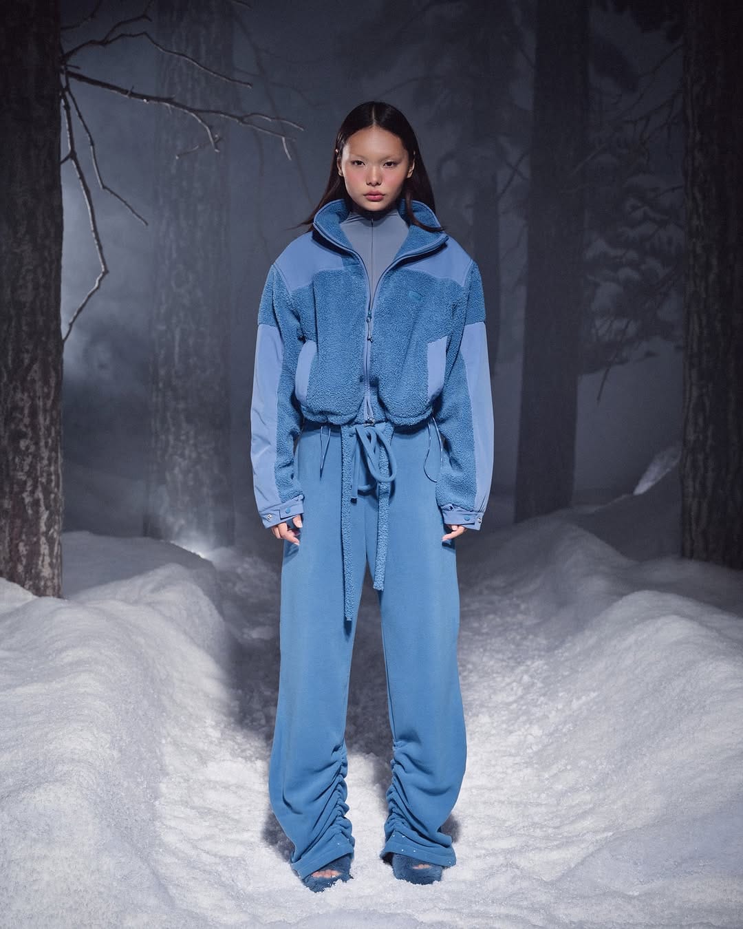 Model standing on a snowy forest path wearing a blue fleece jacket and blue trousers for SKIMS.