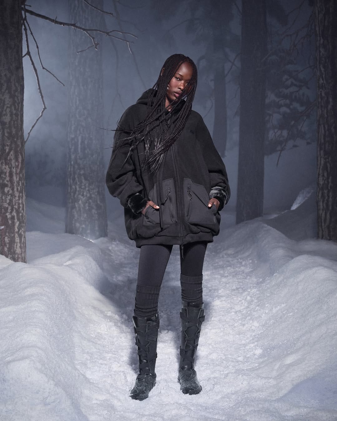Model with long braided hair wearing a black fleece jacket and tall boots on a snowy forest path at night.