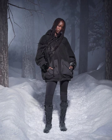 Model with long braided hair wearing a black fleece jacket and tall boots on a snowy forest path at night.