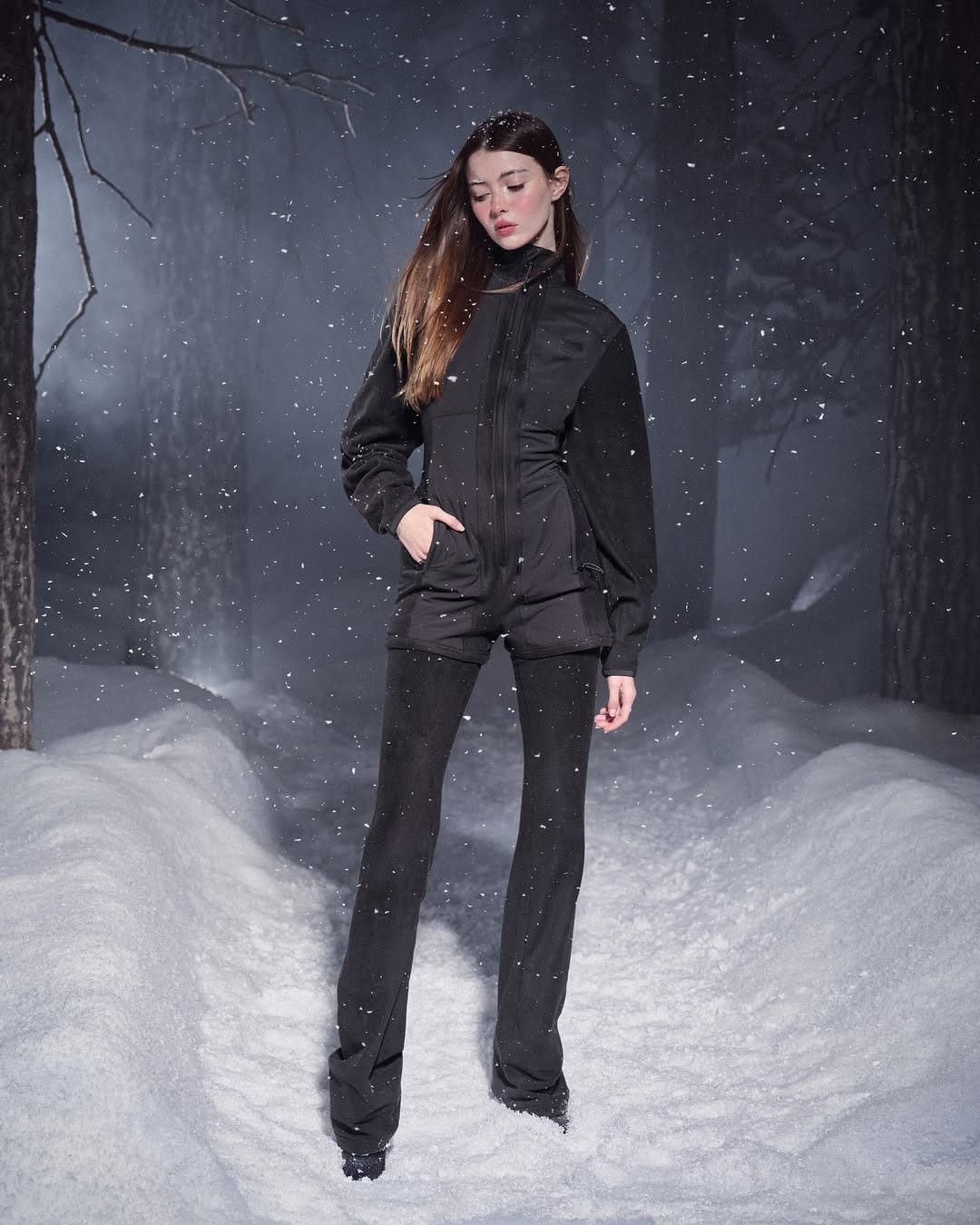 Model wearing a black fleece zip-up jacket and matching trousers standing outdoors in a snowy forest during a snowfall.