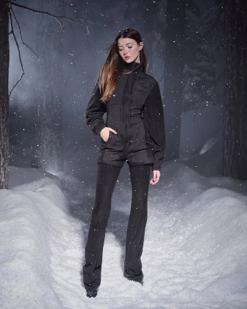 Model wearing a black fleece zip-up jacket and matching trousers standing outdoors in a snowy forest during a snowfall.