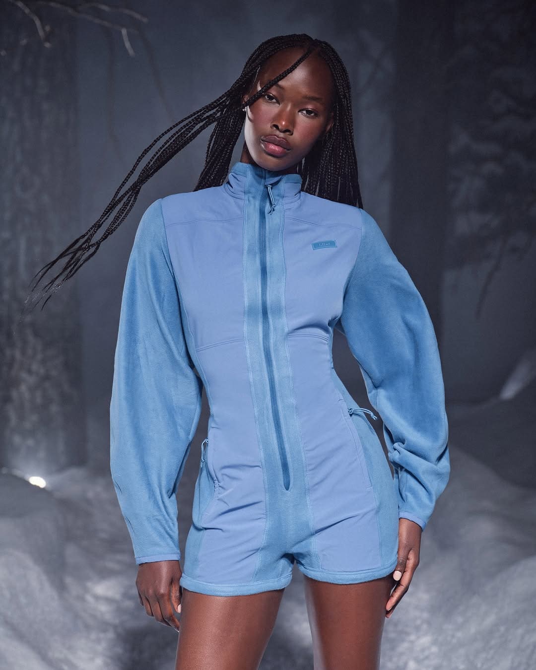 Model with long braided hair wearing a blue fleece jumpsuit standing against a snowy winter background.