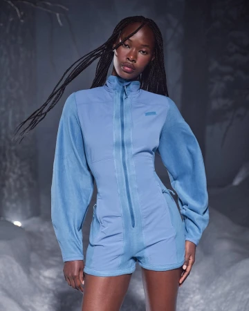 Model with long braided hair wearing a blue fleece jumpsuit standing against a snowy winter background.