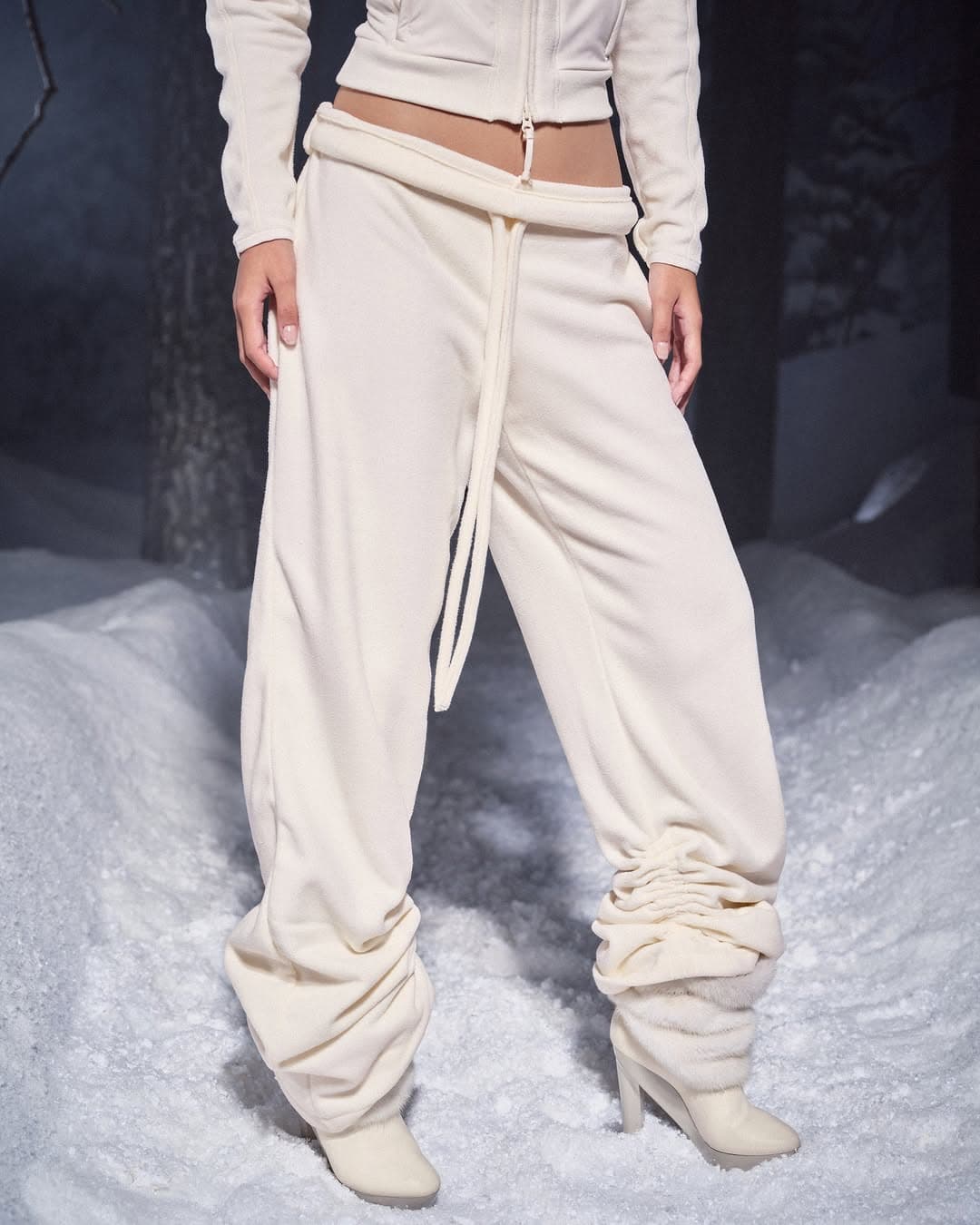 Model wearing cream colored sweatpants and matching zip jacket standing outdoors on snow in a winter setting.