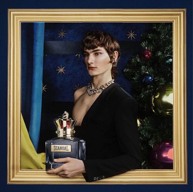 Man wearing a black blazer and silver chain necklace holding a fragrance bottle inside a decorative gold frame