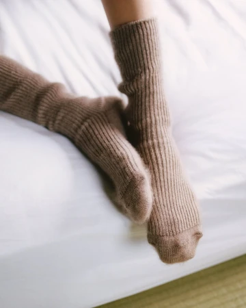 Close-up of legs wearing ribbed brown wool socks resting on a white bed sheet
