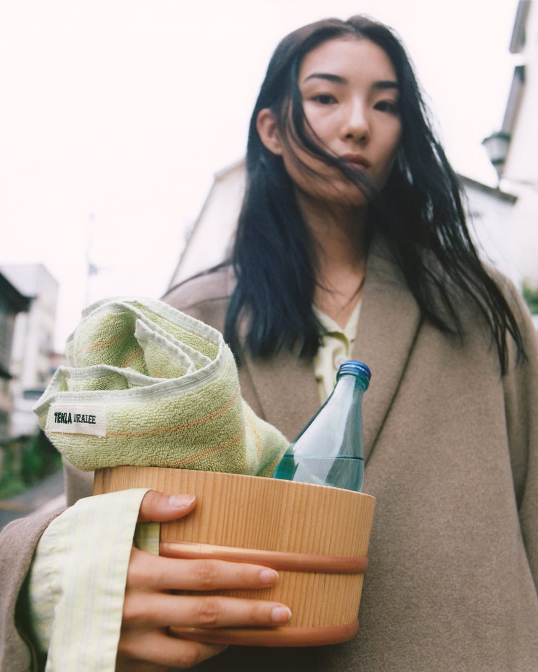 Model holding a wooden bucket with a rolled towel and water bottle in a campaign for Auralee x Tekla