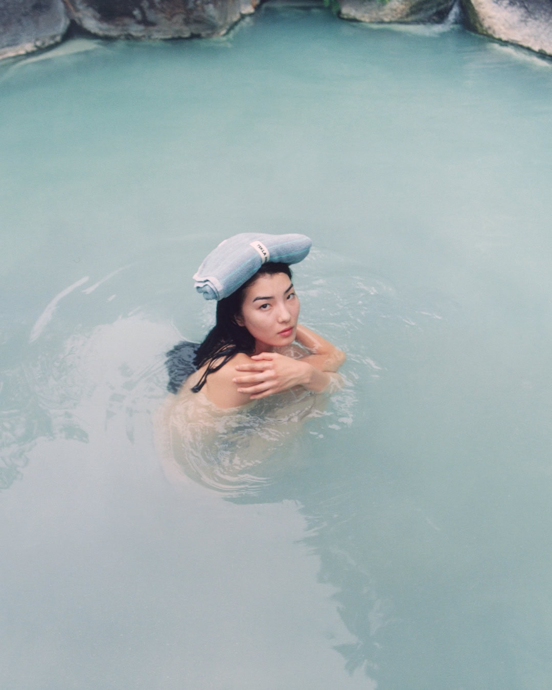 Model in natural hot spring water with a folded towel on her head for the Auralee x Tekla Fall 2024 ad campaign