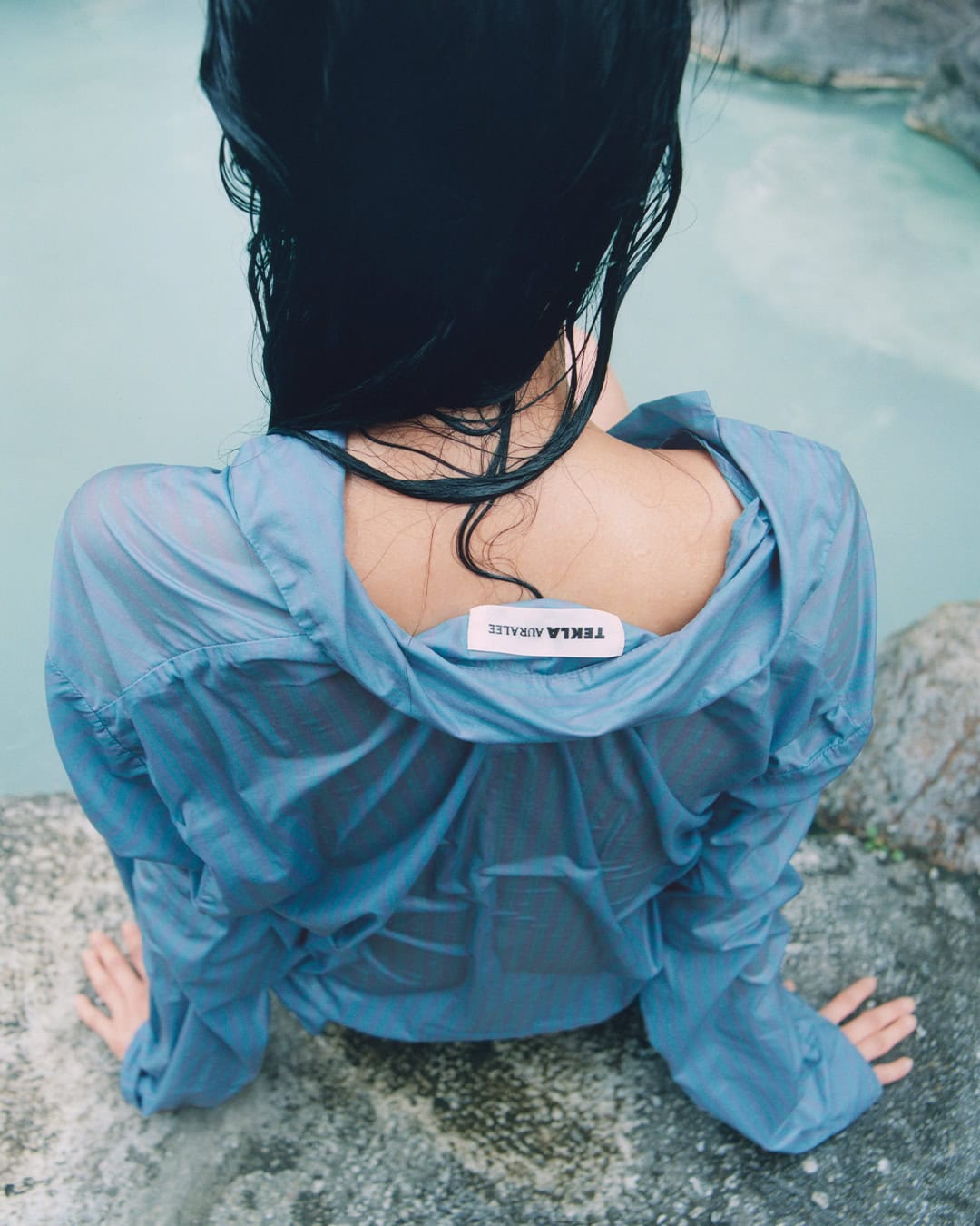 Model with wet black hair wearing a blue sheer shirt sitting on rocky ground by water.