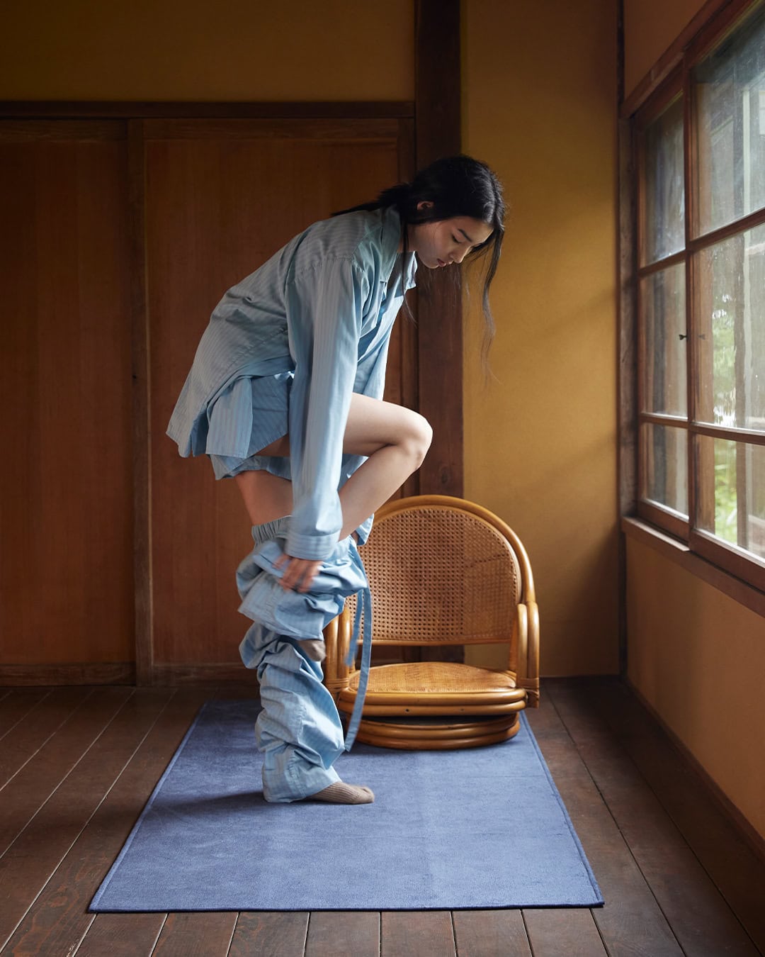Model standing on a blue mat in a wood-paneled room, putting on light blue striped trousers for Auralee x Tekla.