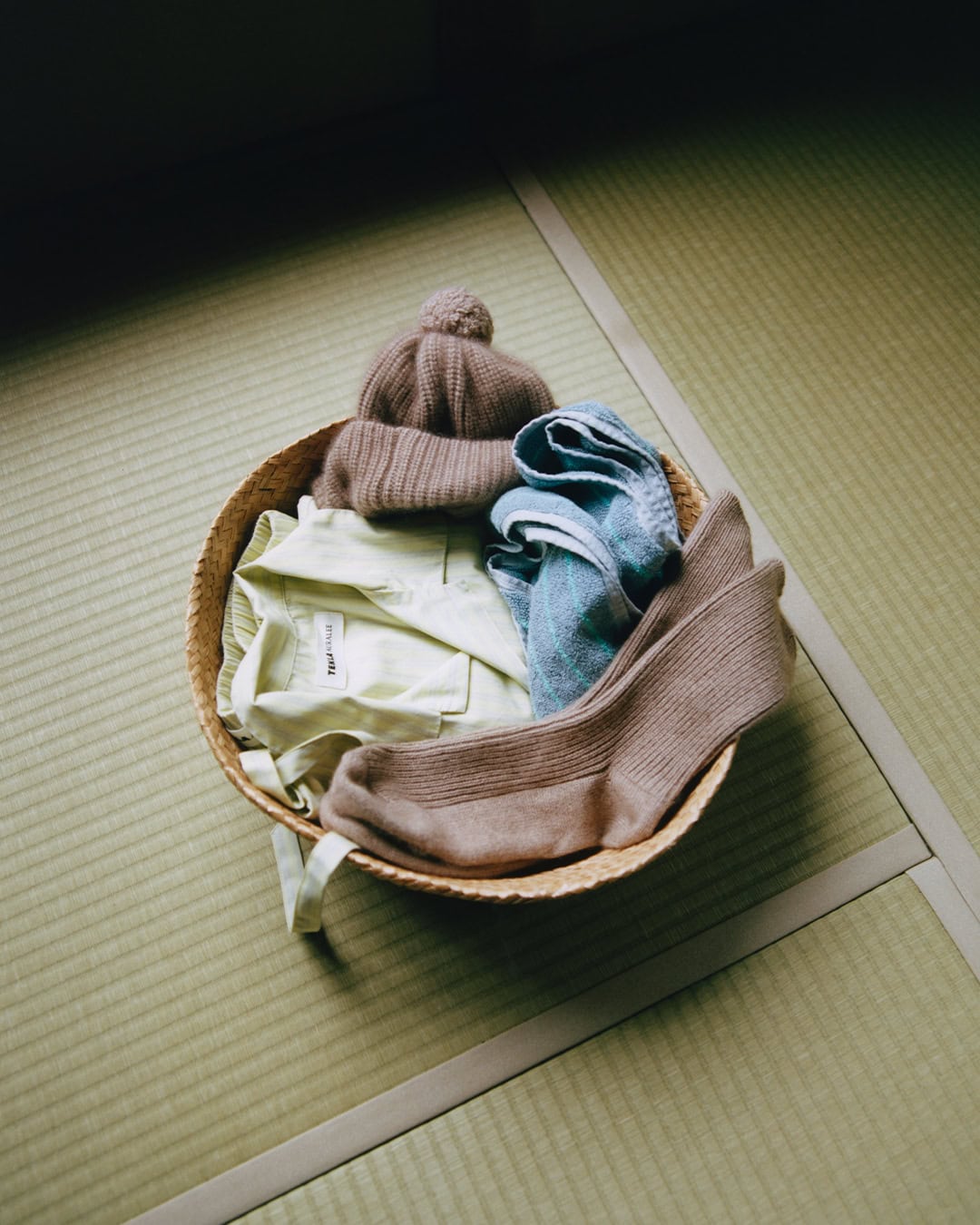 Round woven basket containing a folded pale green shirt, brown knit beanie, blue fabric item, and brown socks on tatami mat flooring.