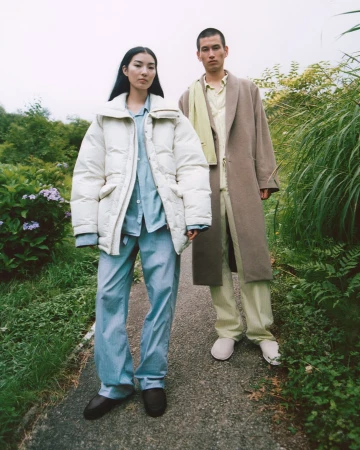Two models standing on an outdoor path wearing winter coats and light apparel for the Auralee x Tekla campaign.