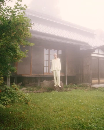 Model standing on a rock in front of a traditional Japanese house shrouded in fog for Auralee x Tekla