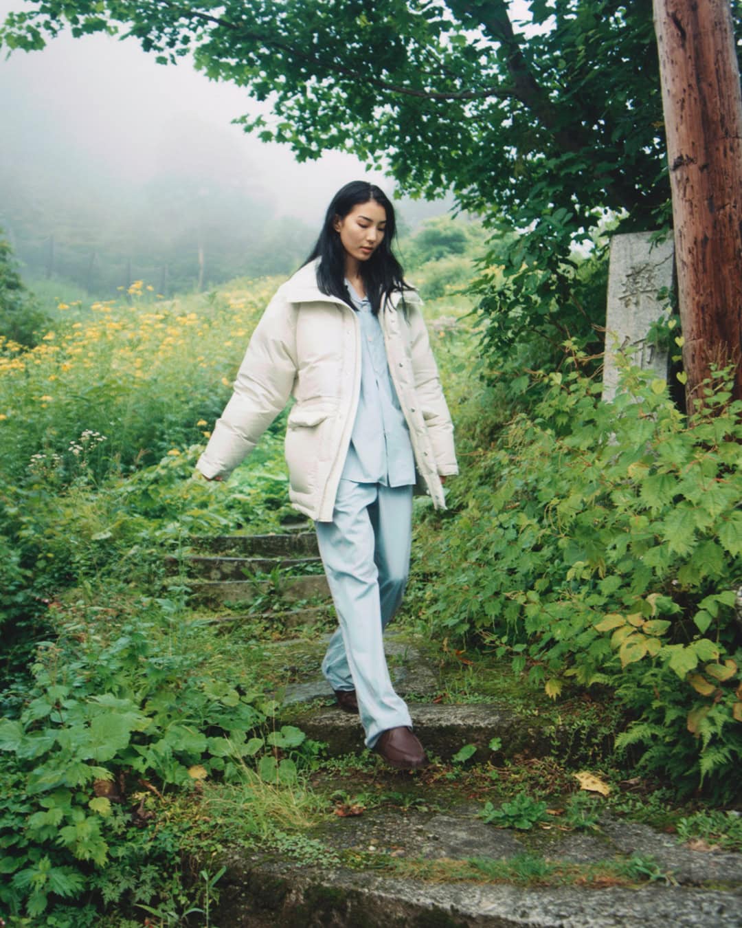 Model walking down a stone path in a green forest wearing a cream puffer jacket and light blue trousers for Auralee x Tekla.