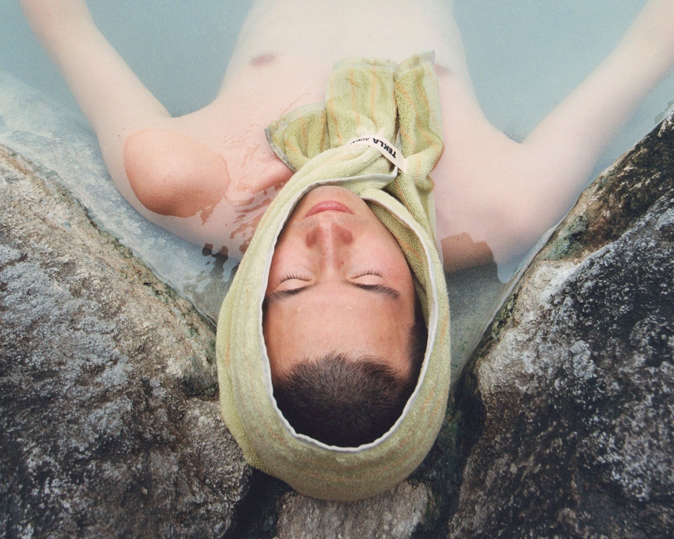Man relaxing in thermal water with a green towel wrapped around his head between rock formations