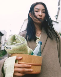 Model holding a wooden bucket with a rolled towel and water bottle in a campaign for Auralee x Tekla