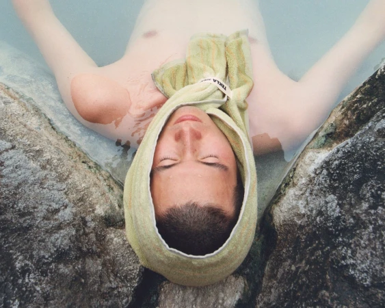 Man relaxing in thermal water with a green towel wrapped around his head between rock formations