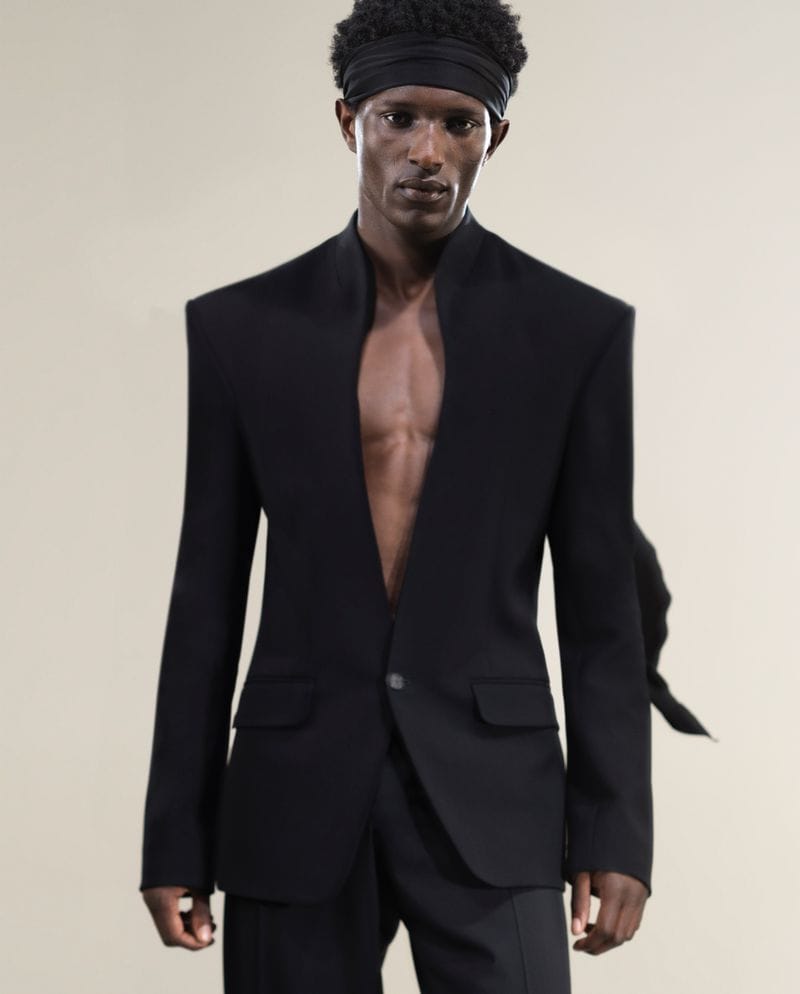 Black man wearing a tailored black suit jacket and headband against a neutral studio background.