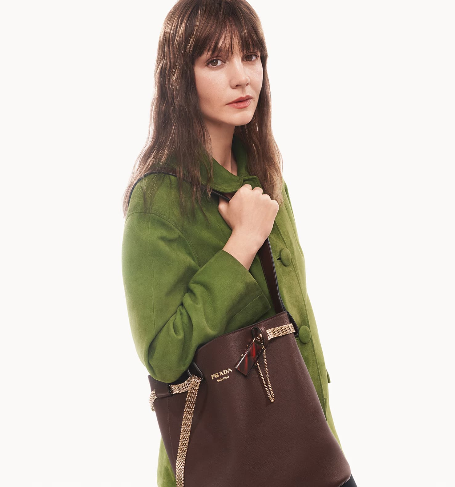 Carey Mulligan wearing a green coat and carrying a brown leather Prada handbag against a white background.