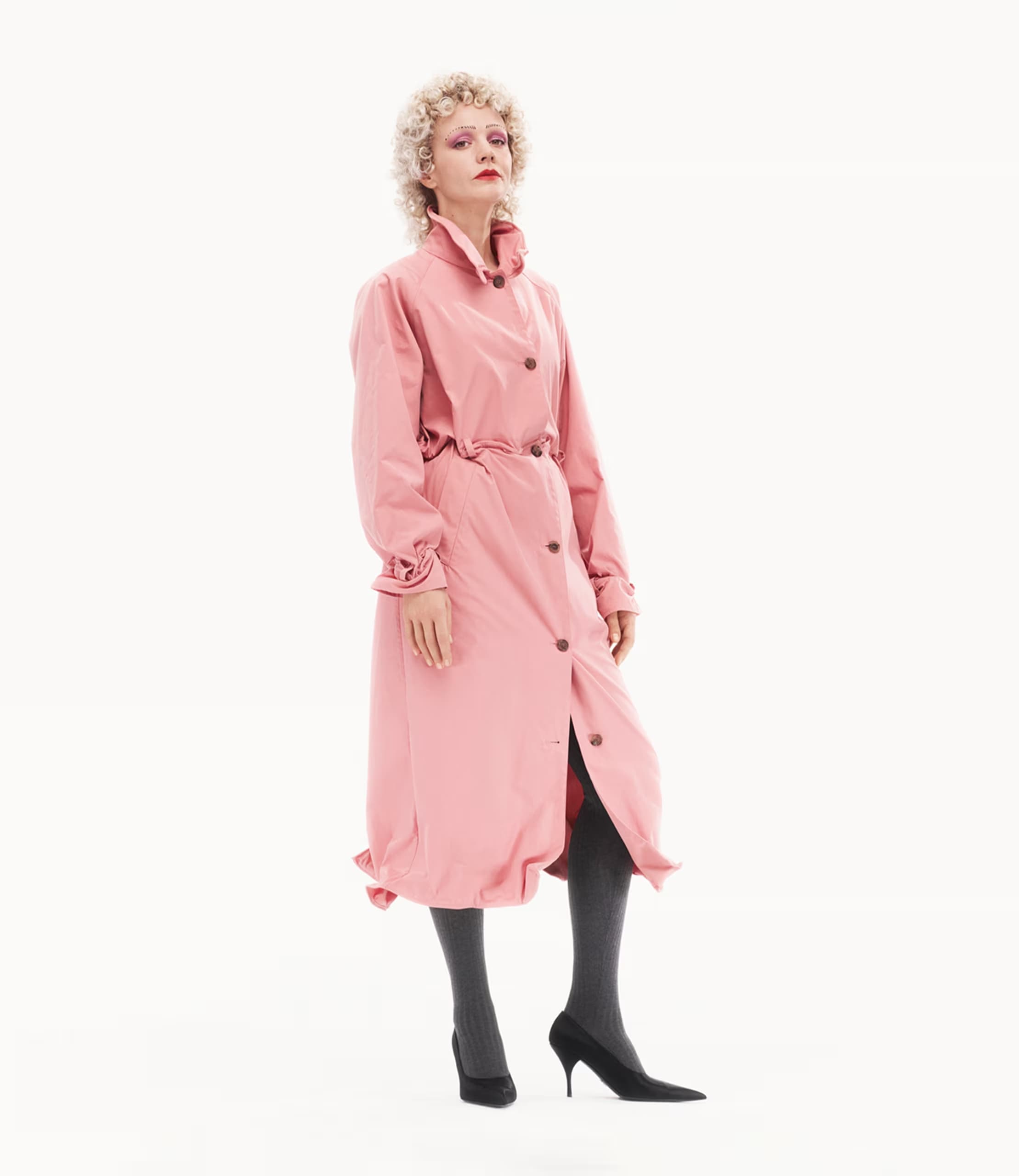Carey Mulligan in a pink trench coat wearing dark tights and heels against a white seamless background for Prada