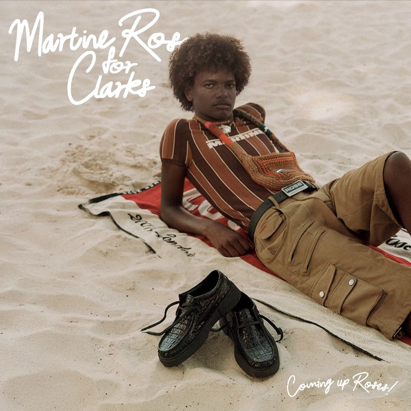 A person poses on a beach wearing a striped top and khaki shorts next to black shoes, with promotional text for Martine Rose and Clarks.