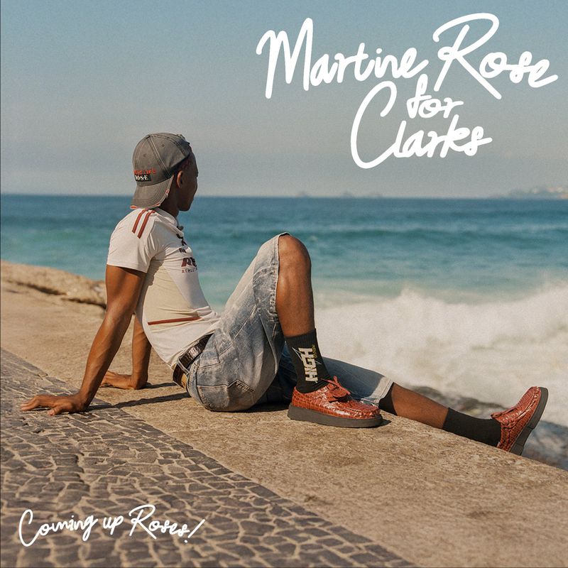 A man in casual clothing sits by the ocean with a sea view backdrop, promoting a fashion collaboration, Martine Rose for Clarks.