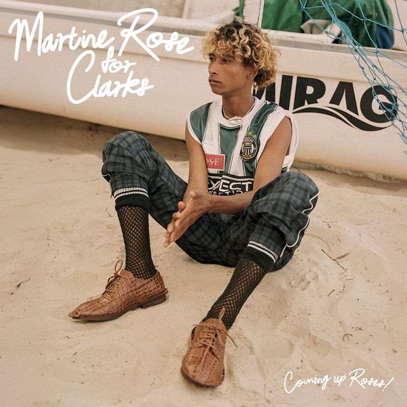 A person sits on a beach in front of a boat wearing a jersey, checkered pants, and brown dress shoes for a fashion editorial.