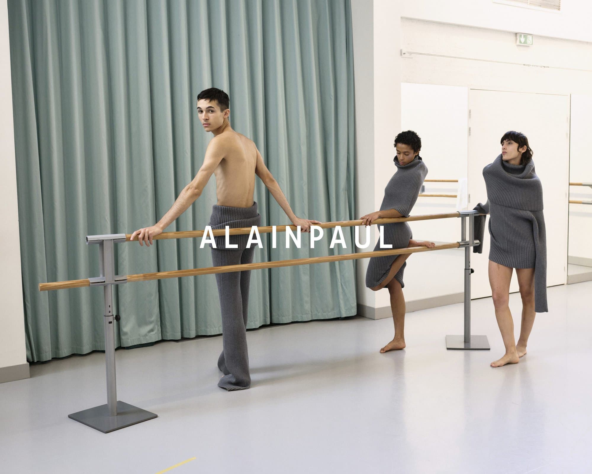 Three models wearing grey knitwear posing by a wooden ballet barre inside a studio with pale floors and teal curtains.