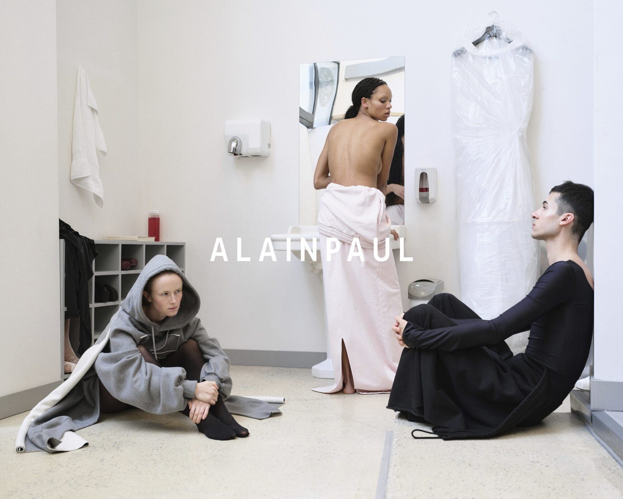 Three models in an interior setting featuring a grey hoodie, pink towel wrap, and black outfit for Alainpaul fall 2024 ad campaign.